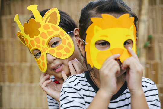顽皮的两个亚洲小女孩喜欢戴着卡通动物面具狮子和长颈鹿坐在一起。面具狂欢节。