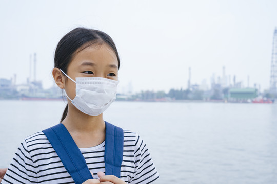 病毒防护概念-戴着防护面具的亚洲小女孩，在雾霾天站在河边。到处都是污染。