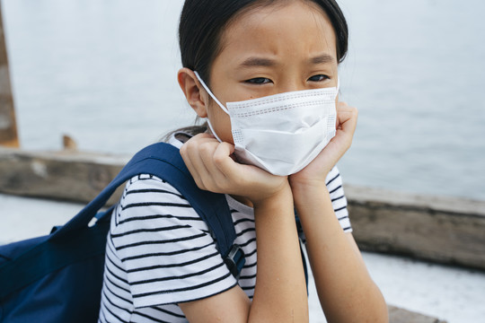 特写：戴着外科口罩的泰国小孩-病毒防护概念-戴着防护口罩的亚洲小女孩，在雾霾天站在河边。到处都是污染