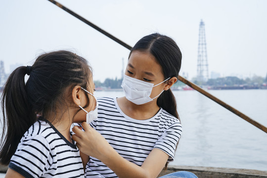 亚洲小孩帮妹妹戴口罩。病毒防护概念-戴着防护面具的亚洲小女孩，在雾霾天站在河边。到处都是污染。