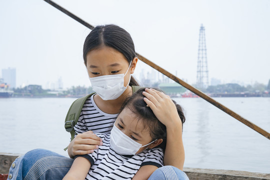 病毒防护概念-戴着防护面具的亚洲小女孩，在雾霾天站在河边。到处都是污染。泰国大姐姐抱着小妹妹。仁慈。
