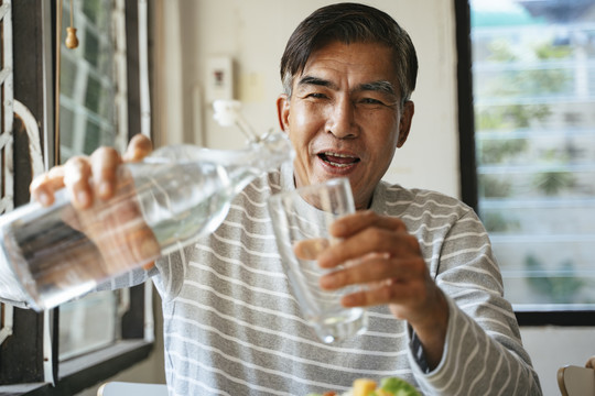 老人手里拿着玻璃杯和一瓶水，他将饮用水倒进玻璃杯里。