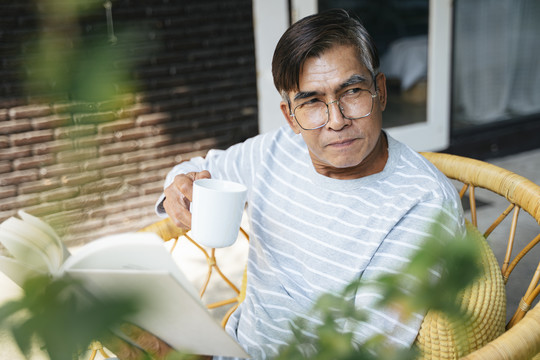 快乐的老人坐在院子里的柳条椅上，手里拿着书和白杯子。