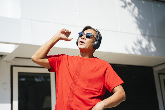在阳光明媚的日子里，穿着红色t恤衫的老人在家门口欣赏耳机里的音乐，跳舞。