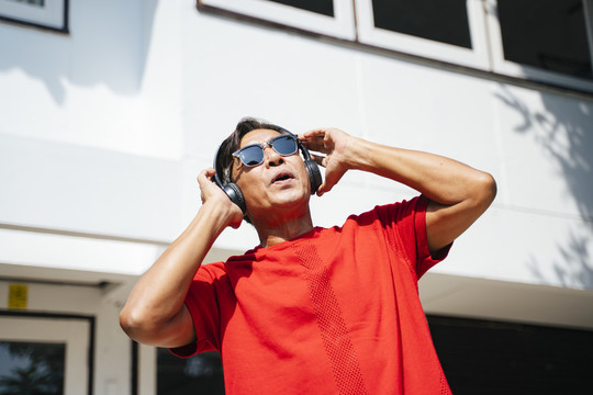 在阳光明媚的日子里，穿着红色t恤衫的老人在家门口欣赏耳机里的音乐，跳舞。