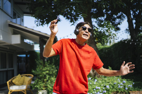 穿着红色t恤的老人手持智能手机，戴着真正的无线耳机，在家里的阳光下翩翩起舞。