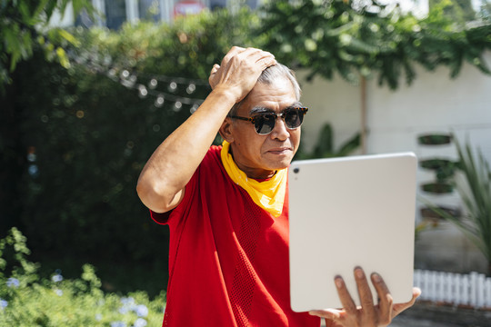 身穿红色t恤、戴着墨镜的老人看着平板电脑上的倒影，对自己的外表感到自豪。