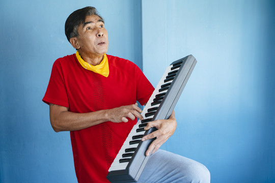 穿着红色t恤和黄色围巾的老人在蓝色的房间里玩电子键盘。