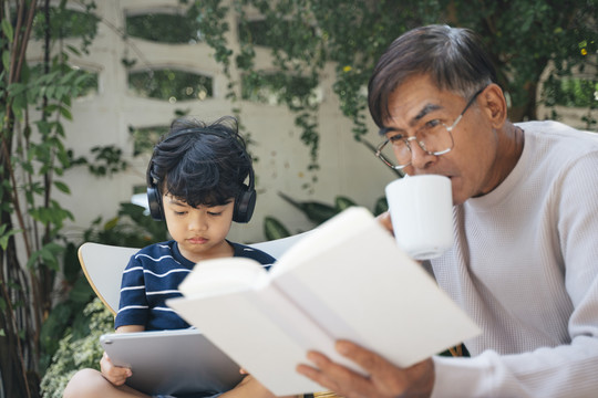 小男孩戴着耳机听音乐，读平板电脑上的电子书，而老人喝着咖啡，读着书。