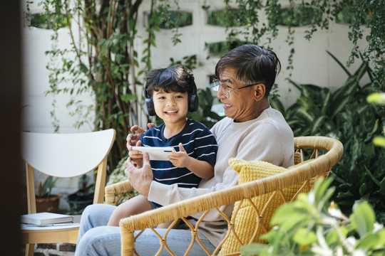 小男孩和爷爷坐在后院的柳条椅上，戴着无线耳机，用智能手机玩游戏。