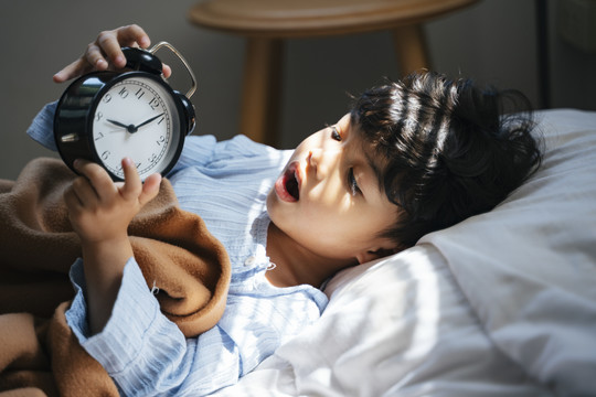 小男孩白天躺在床上睡觉，对古董闹钟很感兴趣。