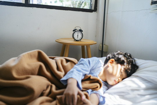 古董闹钟放在床旁边的木桌上，上面放着中午睡觉的男孩。