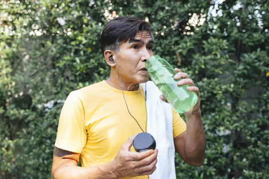 穿黄色t恤的老人慢跑后休息一下，他喝下绿色运动瓶里的水，用白毛巾擦汗。
