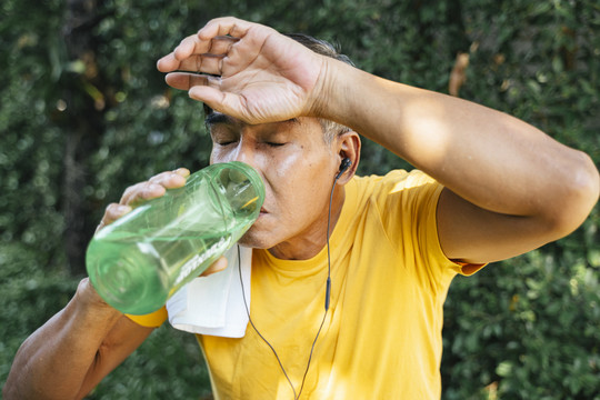 穿黄色t恤的老人慢跑后休息一下，他喝下绿色运动瓶里的水，用白毛巾擦汗。