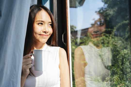 在夏日的阳光下，美丽的长发女人站在带窗帘的玻璃窗前。