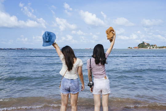 两个女孩在海边玩得很开心，她们在海边旅行，向天空高举帽子。