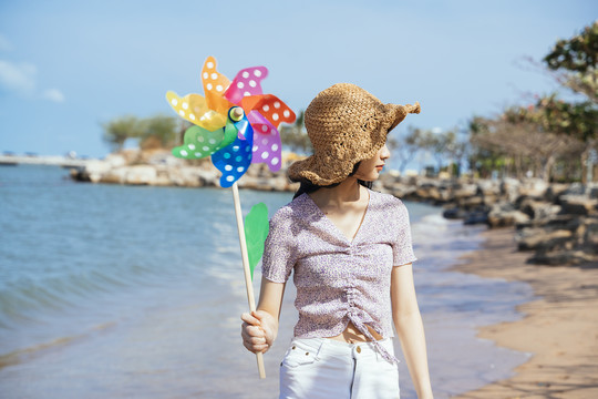 可爱的女孩戴着棕色的帽子拿着五颜六色的风车在沙滩上散步在大风天。