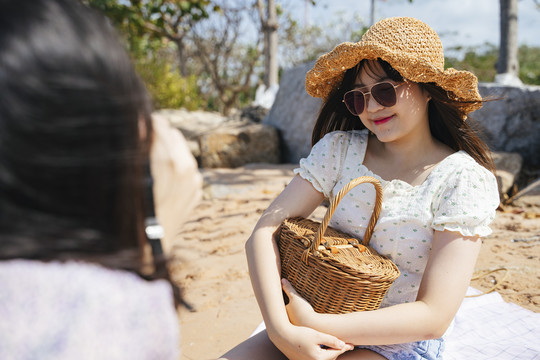 戴着棕色帽子和太阳镜的长发女孩坐在那里，抱着木篮，她的朋友在给她拍照。