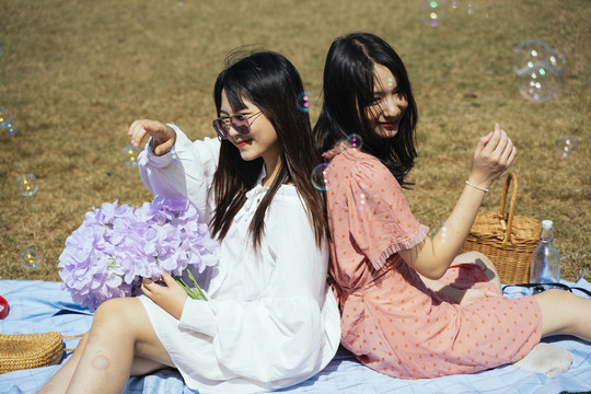 两个女孩背靠背地坐在地上的垫子上，玩着泡泡和紫花。