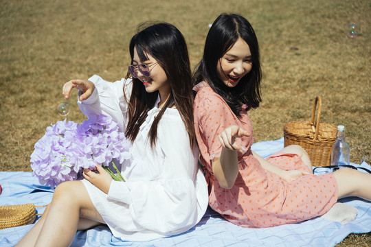 两个女孩背靠背地坐在地上的垫子上，玩着泡泡和紫花。