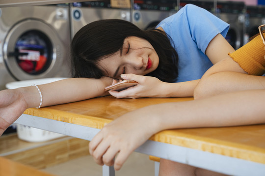 两个女孩在自助洗衣店等衣服时睡在木桌旁。