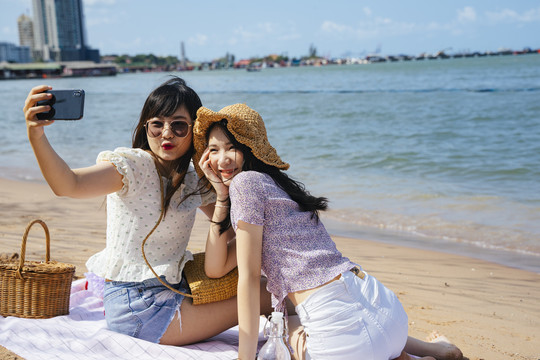 阳光明媚的日子里，两个女孩坐在海边的沙滩上，一起自拍，做鸭脸。