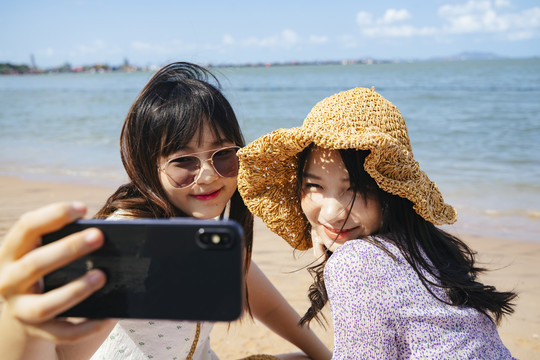 阳光明媚的日子里，两个女孩坐在海边的沙滩上，一起自拍，做鸭脸。