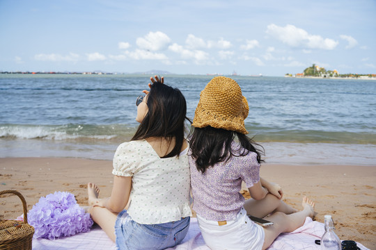 风大的时候，两个女孩背靠背地坐在沙滩上的垫子上，享受周末的快乐。