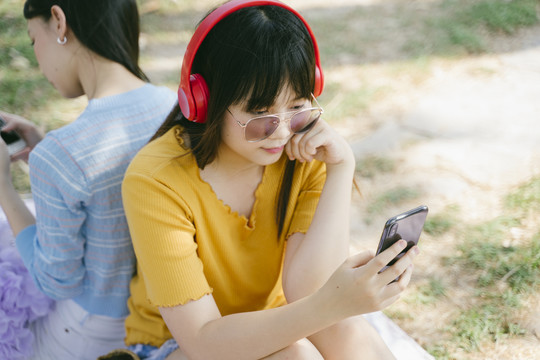 两个女孩背靠背地坐在地板上，听着智能手机应用程序里的歌曲，在社交媒体上旅行。