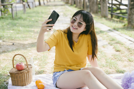 一个穿着黄色t恤的女人在野餐时戴着墨镜坐在垫子上自拍。