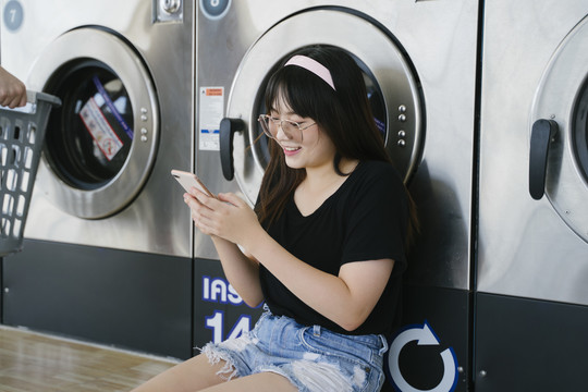 戴眼镜的长发女孩在等待洗衣机时使用智能手机。