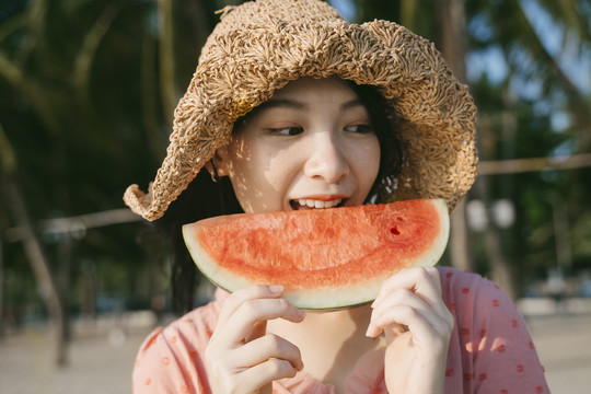 一个戴棕色帽子的女孩站在沙滩上吃西瓜。