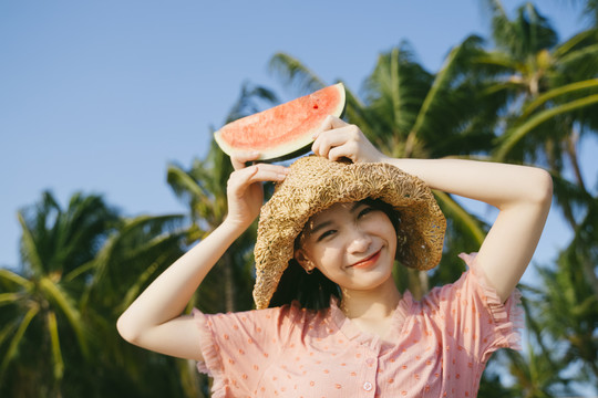 可爱的女孩把西瓜放在头上，微笑着站在沙滩上。