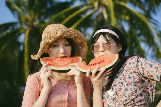 傍晚日落时，两个女孩站在沙滩上吃西瓜。
