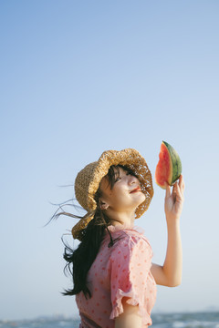 戴着棕色帽子的可爱女孩抬起头上的西瓜，在蓝天下微笑。