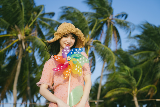 风和日丽的沙滩上，戴着棕色帽子的可爱女孩手持五颜六色的风车在椰子树前。