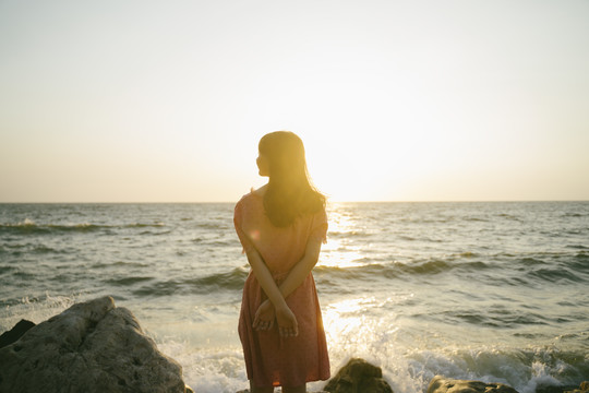 长发女孩站在海边的岩石上，悠闲地看着夕阳。