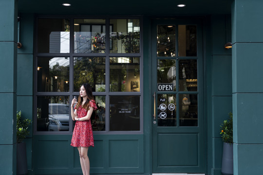 一位身穿红色连衣裙的亚洲年轻女子在绿色咖啡厅门前端着一杯咖啡。