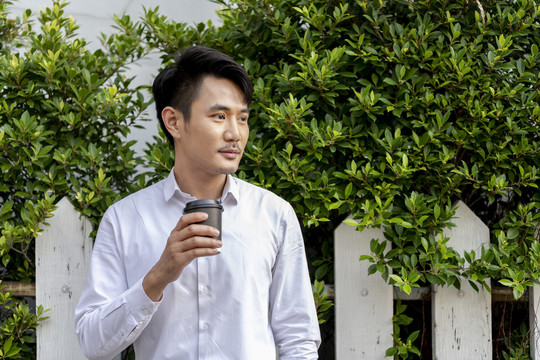 穿着白衬衫的泰国成年商人在户外拿着一杯咖啡，背景是大自然。