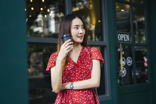 身着红色连衣裙的亚洲年轻女子在咖啡厅门前享用热咖啡。