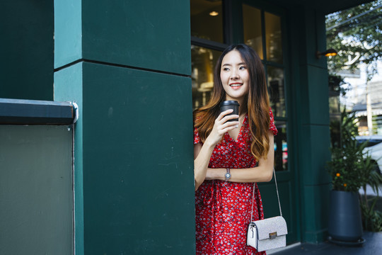 在户外咖啡馆里，一位身着红色连衣裙、手持咖啡的快乐的亚洲年轻女子。