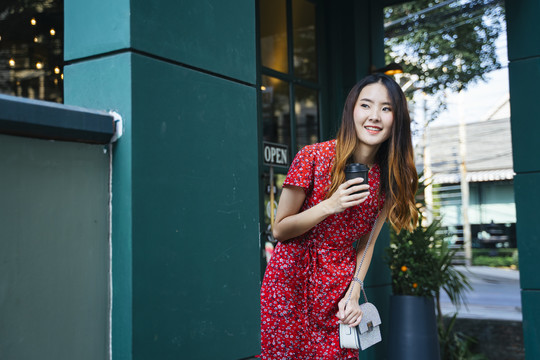 在户外咖啡馆里，一位身着红色连衣裙、手持咖啡的快乐的亚洲年轻女子。