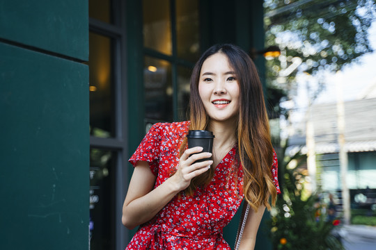 在户外咖啡馆里，一位身着红色连衣裙、手持咖啡的快乐的亚洲年轻女子。