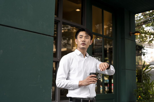 成年亚泰商人穿着白衬衫，端着一杯咖啡，看着手边的手表，看时间去了。