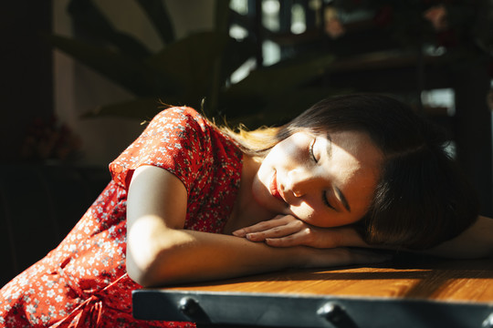 一位身穿红色连衣裙的泰国亚裔年轻女子在屋内的阳光下睡在桌子上。