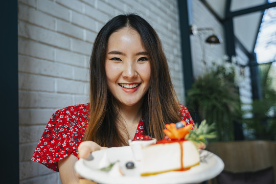 身穿红色连衣裙的泰国亚裔年轻女子，手里拿着白色盘子，盘子上放着奶酪蛋糕。快乐的表情。