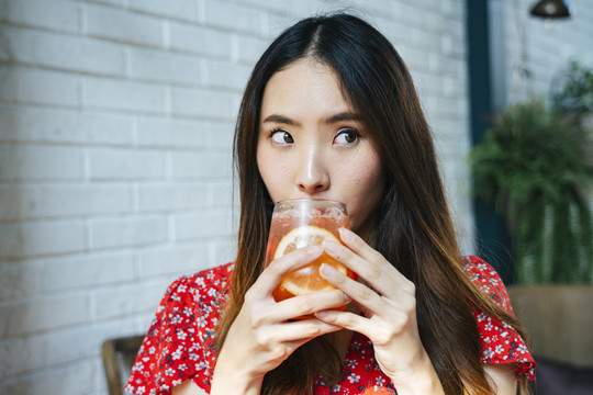 特写身着红色连衣裙的泰国亚裔年轻女子，双手捧着冰镇果汁，一边喝酒，一边啜饮。
