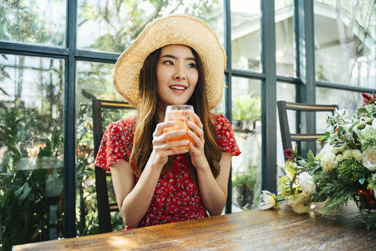 在绿色咖啡厅，一位身穿红色连衣裙、头戴草帽、喝着冰镇果汁的泰国亚裔年轻女子。