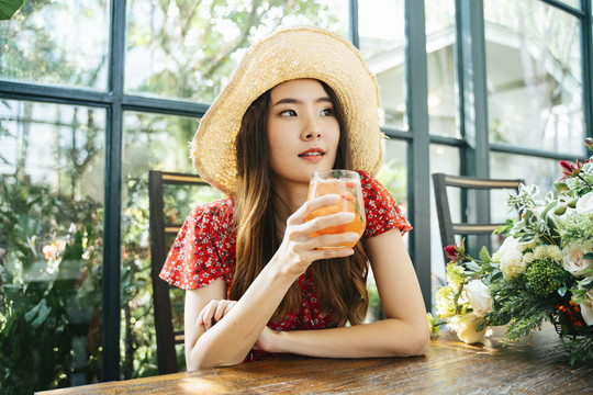 在绿色咖啡厅，一位身穿红色连衣裙、头戴草帽、喝着冰镇果汁的泰国亚裔年轻女子。