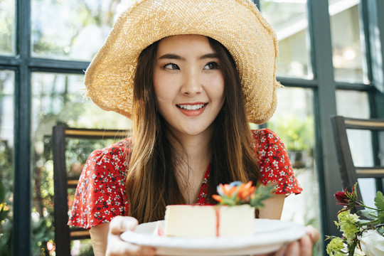 特写镜头：身穿红色连衣裙、戴着帽子、拿着芝士蛋糕盘的泰国亚裔年轻女子。甜点爱好者。